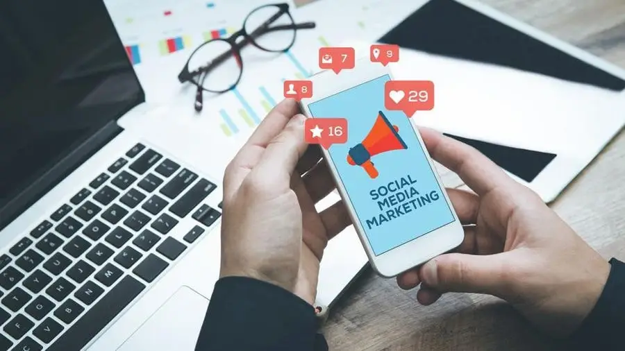 Person holding smartphone displaying 'Social Media Marketing' with notification icons, next to laptop and glasses on desk.